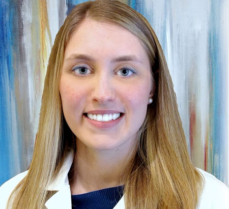 The image shows a woman wearing a white lab coat, smiling at the camera, with blonde hair and blue eyes. She has a professional appearance and is likely associated with a medical or scientific field due to her attire.