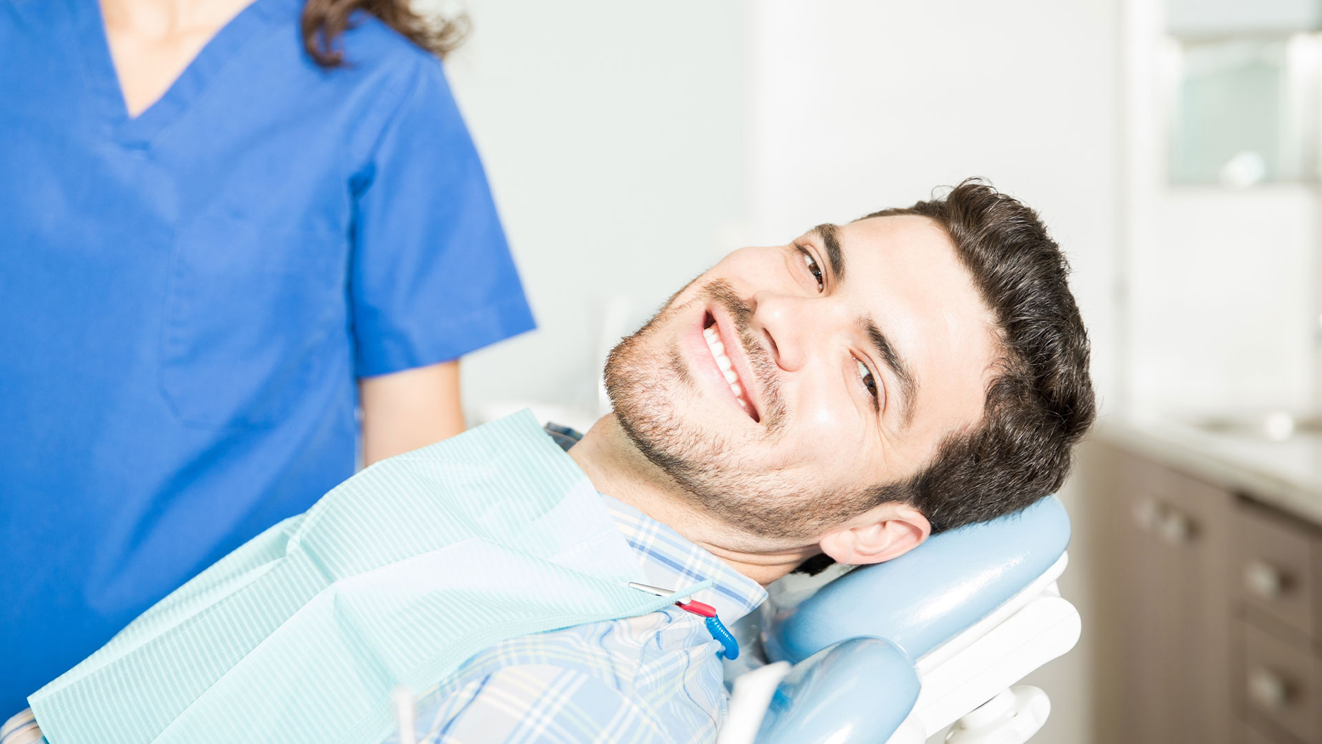 The image features a man sitting in a dental chair with a bright smile, wearing a blue shirt, receiving dental care from a female dentist who is standing behind him and looking at him with a warm expression.