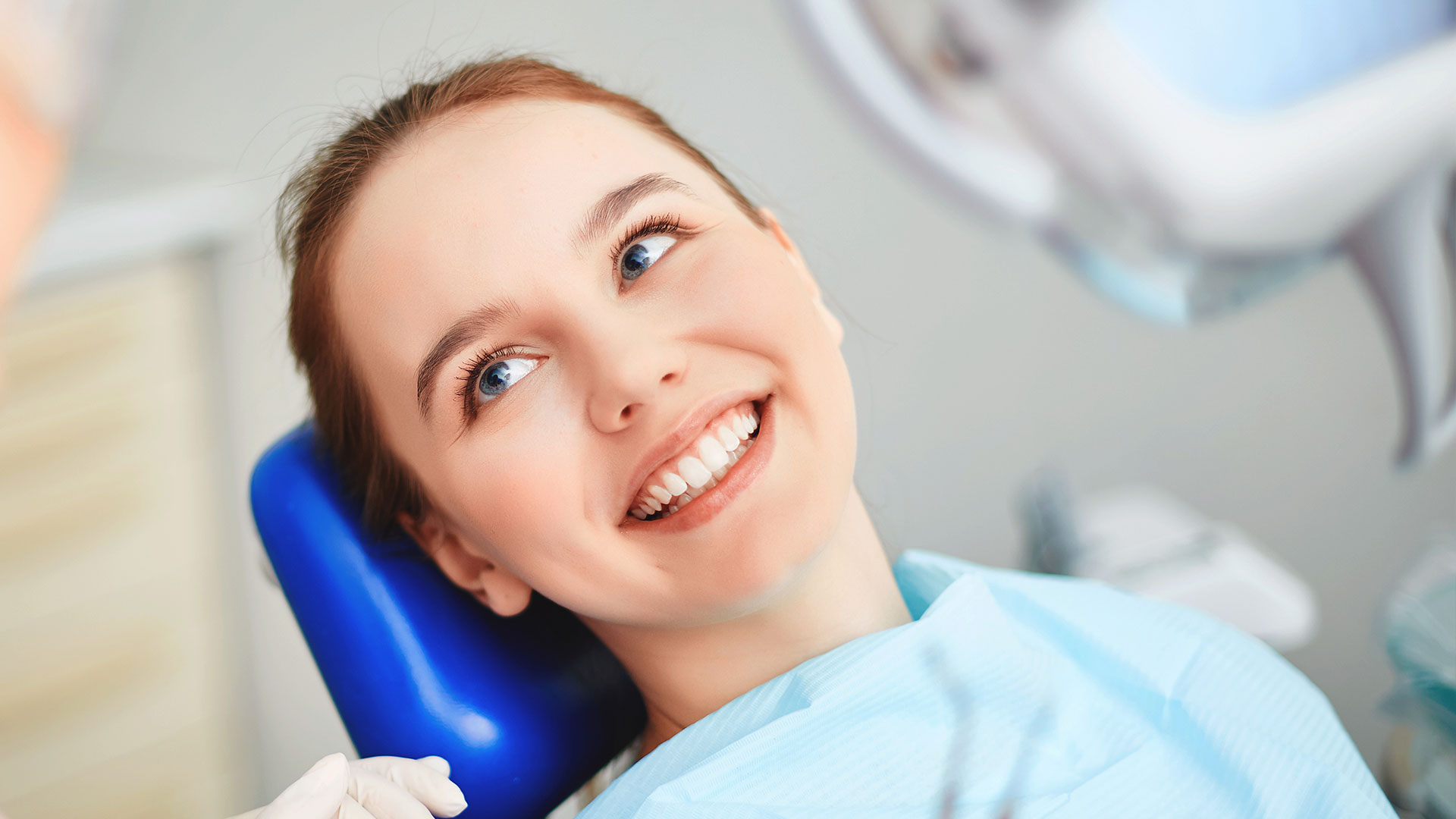 A young woman with a broad smile is seated in a dental chair, looking directly at the camera, while a dentist works on her teeth.