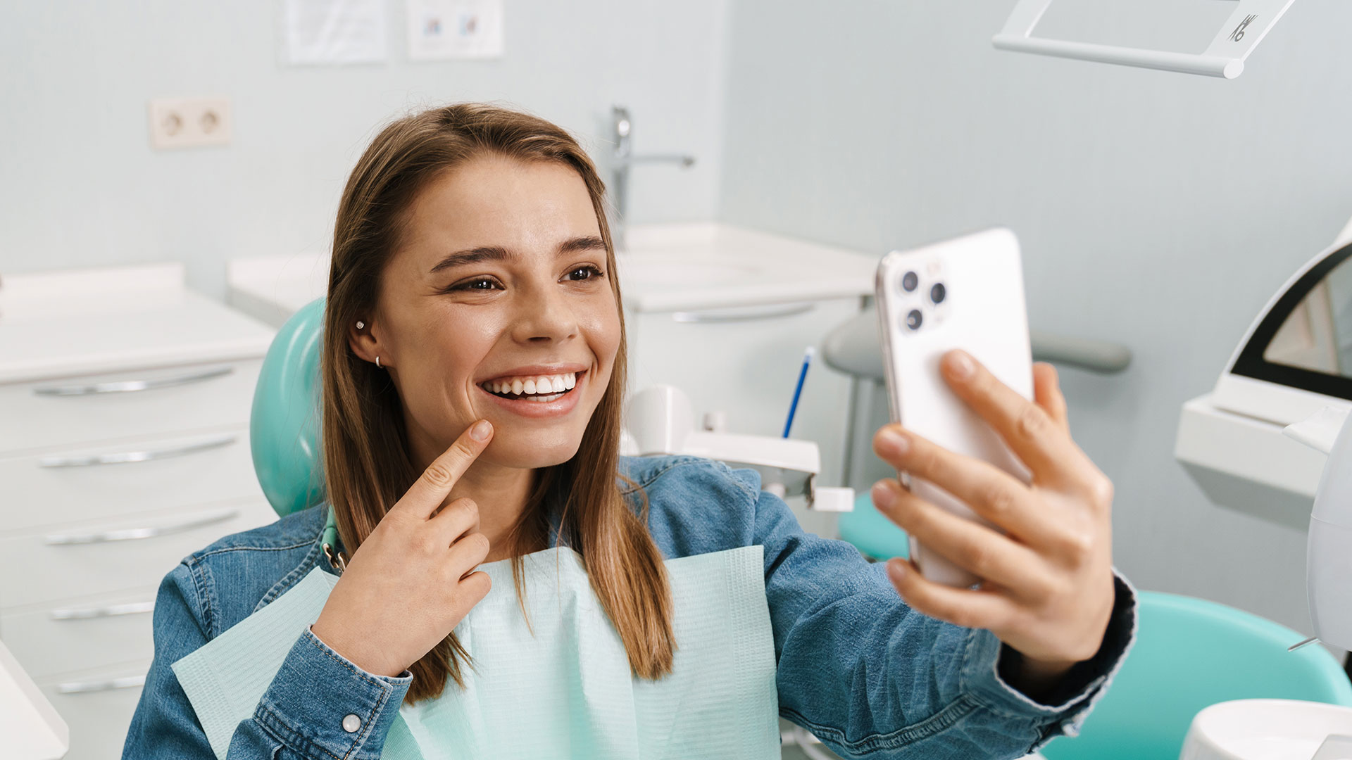 A young woman with a bright smile holds up her phone, taking a selfie in front of a dental office setting.