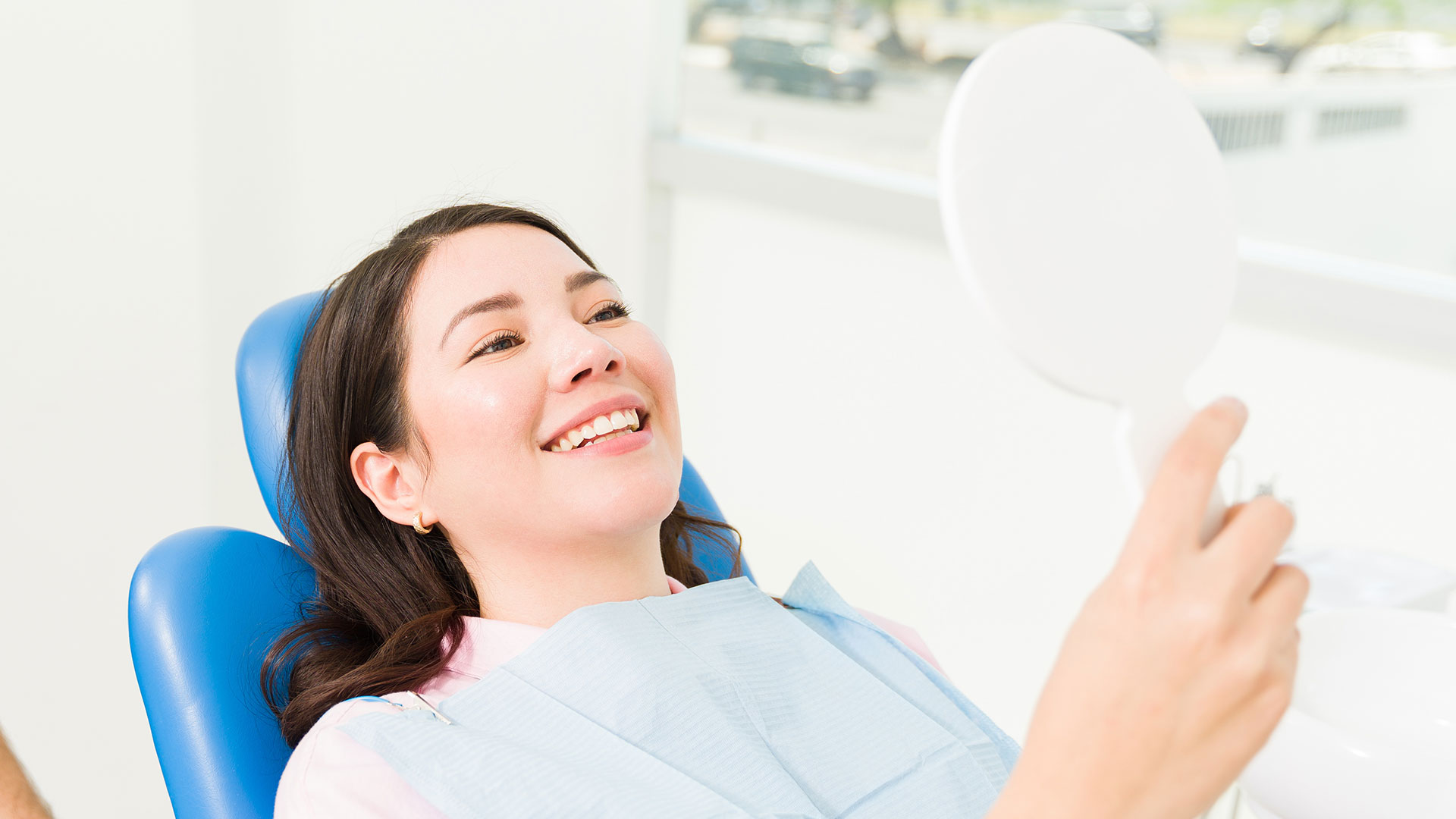 A woman is sitting in a dental chair, smiling, with a mirror in front of her, while a person takes a selfie.