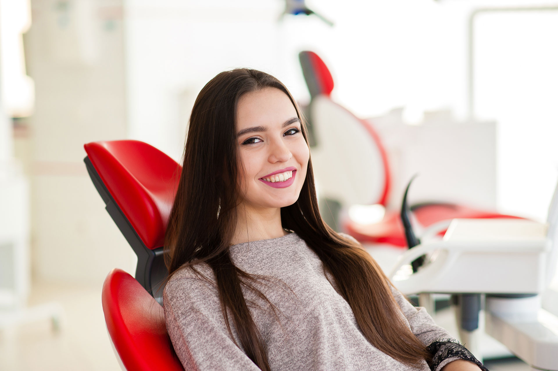 A young woman with long hair sitting in a dental chair, smiling at the camera.