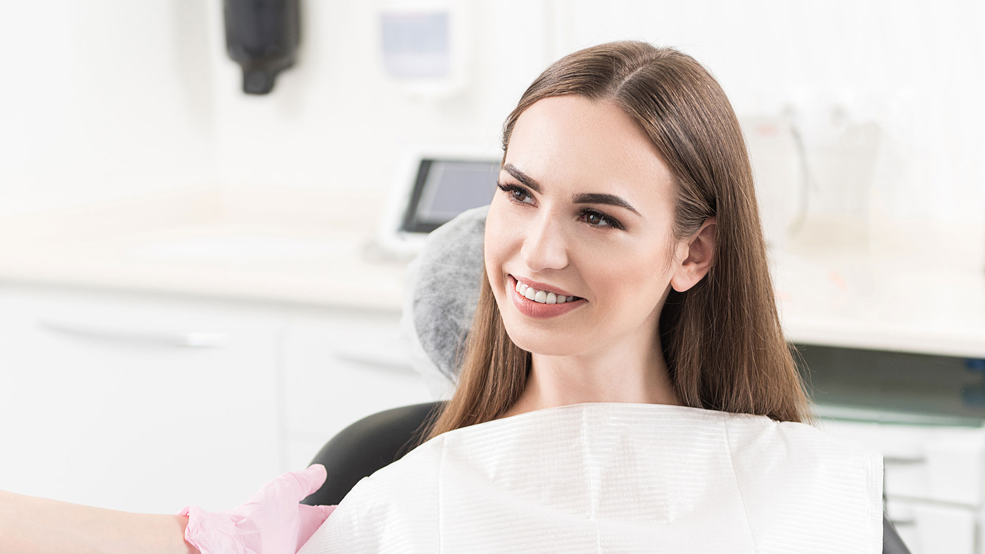 The image shows a smiling woman sitting in a dental chair, with a dentist s office setting visible behind her.