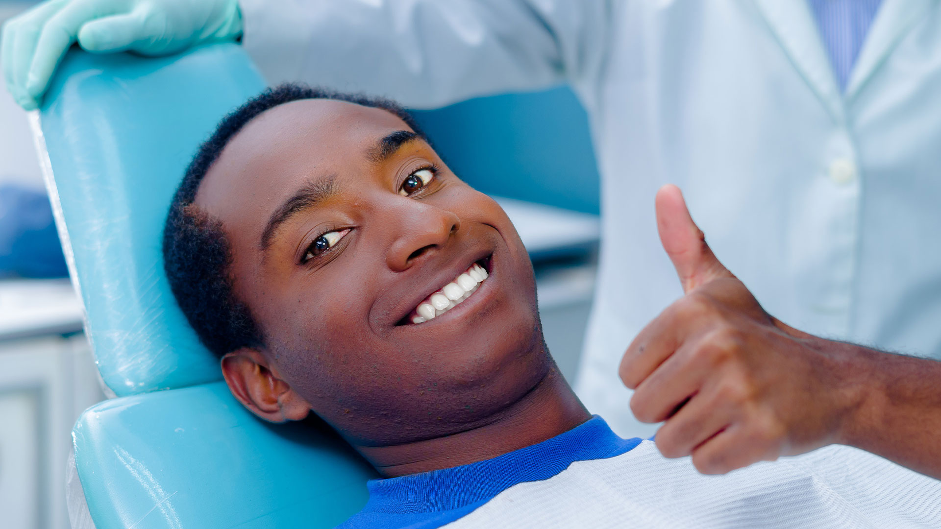 The image shows a dental professional giving a thumbs-up gesture while wearing protective gloves and sitting in a dental chair, with a patient in the background receiving dental care.