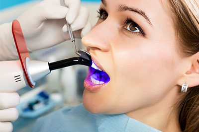 An image depicts a woman receiving dental care, with her mouth open wide, showing a device that appears to be an oral scanner or 3D printer used for dental procedures, while she wears protective eyewear and is seated in a dental chair.