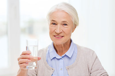 The image shows an elderly woman holding a glass of water with a smile on her face, suggesting she might be promoting hydration or health.