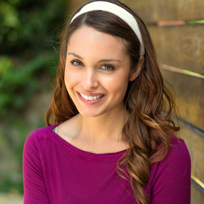 A young woman with long dark hair, wearing a purple top, stands against a wooden fence, smiling at the camera.