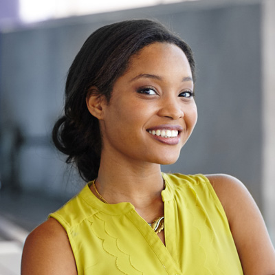 The image shows a smiling woman wearing a yellow top and standing against a wall with a window behind her.