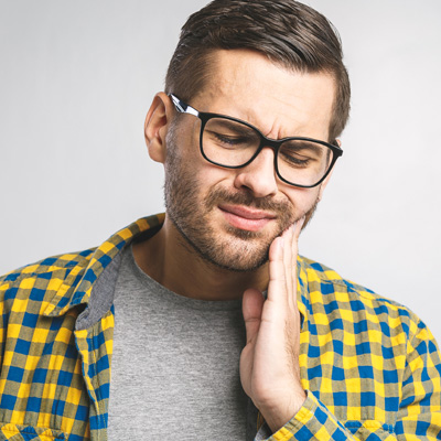 A man with glasses and a beard, wearing a yellow plaid shirt, is seen holding his chin with his hand, possibly displaying discomfort or concern.