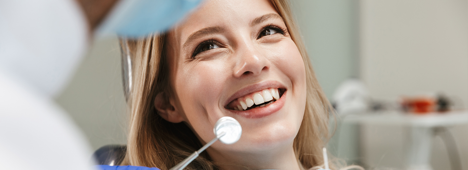 A smiling woman seated in front of a dental chair, looking at a camera, with a dental professional standing behind her, wearing protective gear.