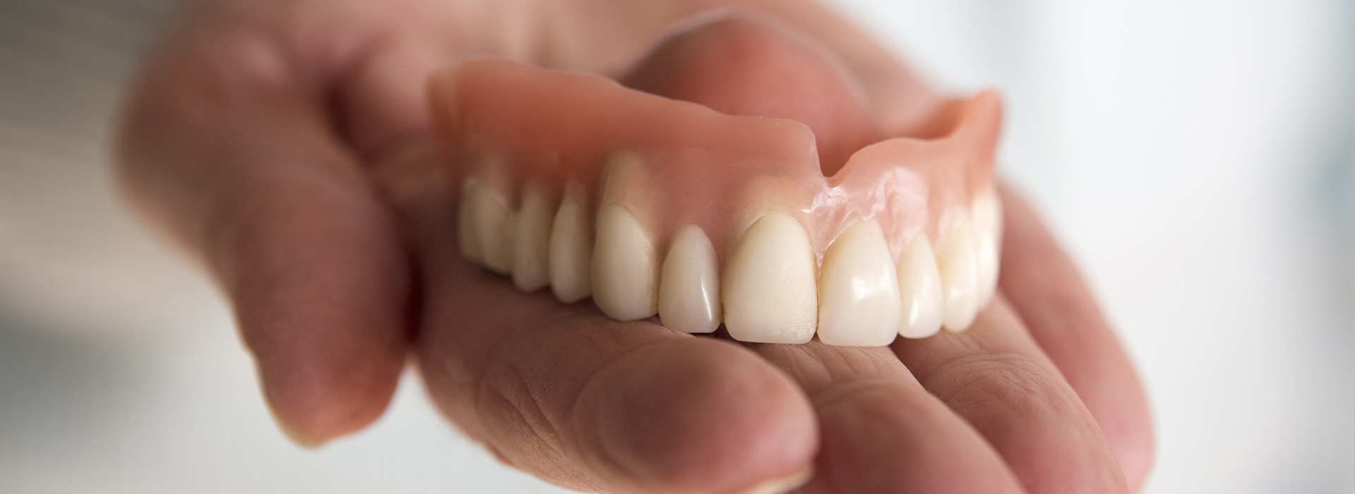 The image shows a person holding an upper denture with artificial teeth against a blurred background.