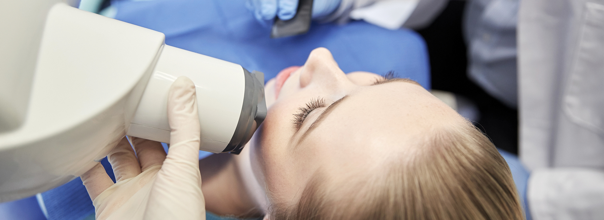 An individual receiving a dental treatment with a dental drill being used on their teeth by a dentist wearing protective gloves and a surgical mask.