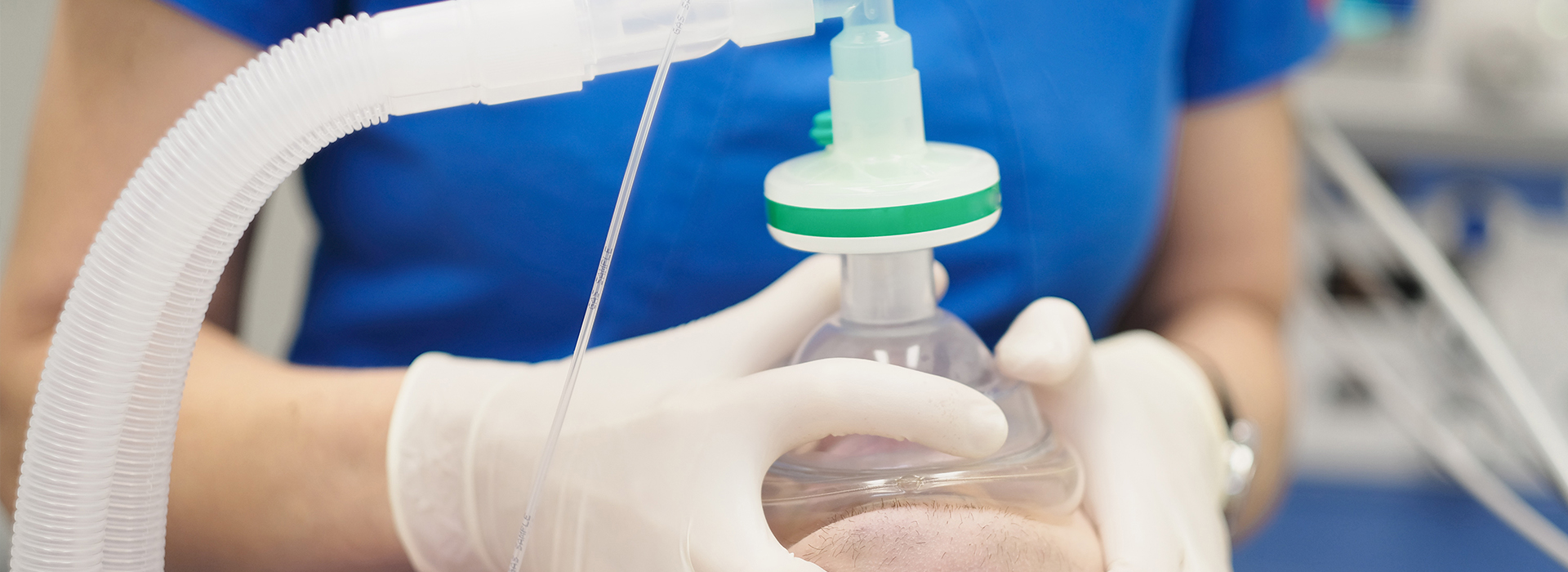 The image shows a person wearing gloves and a blue shirt, handling a clear plastic bottle with a green cap, which appears to be connected to a tube system, possibly for medical purposes.