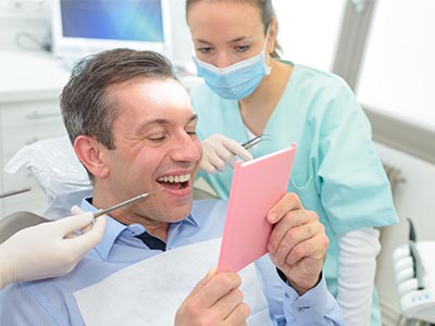 The image shows a man sitting in a dental chair with an open cardboard box in front of him, looking surprised or amused, while a woman wearing medical scrubs stands behind him holding a tablet.