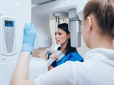 A woman stands next to a large, advanced medical imaging machine, possibly an MRI, while another person appears to be operating or monitoring it. They are both wearing blue scrubs and white gloves, indicating a clinical setting.