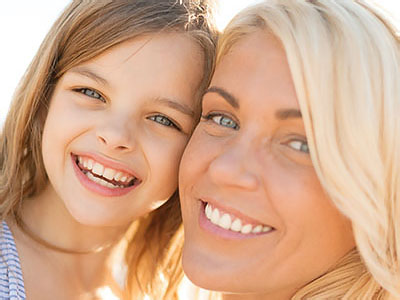 The image shows a woman and a young girl smiling at the camera, with the woman appearing to be a mother and the child her daughter, both enjoying a sunny day outdoors.