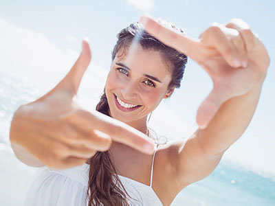 A woman with long hair smiles while holding her hand up to take a selfie against a bright sky background.